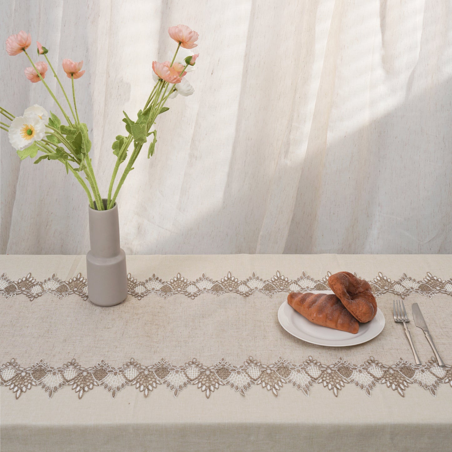 Table setting with a vase of flowers, bread, and cutlery on a tablecloth with lace trim.