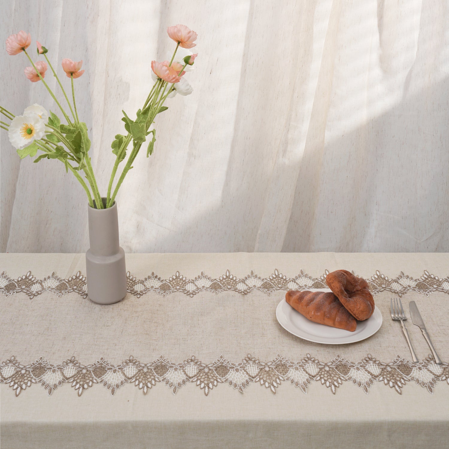 Table setting with a vase of flowers, bread, and cutlery on a tablecloth with lace trim.